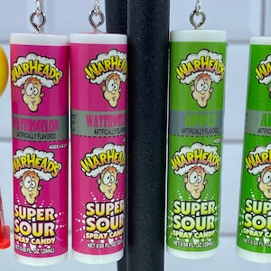 May include: A set of six pairs of earrings featuring miniature candy-themed charms. Two pairs feature yellow smiley faces with hearts for eyes. The other four pairs feature miniature Warheads candy tubes in pink, green, and blue with different flavors: Watermelon, Apple, and Raspberry.