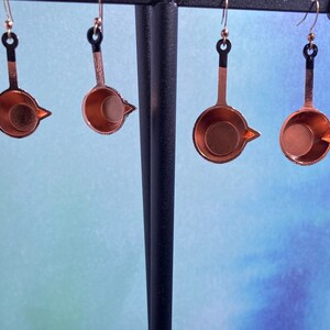 May include: Four pairs of copper-colored earrings shaped like small pots with black handles. The earrings are hanging from a black display stand against a blue and white background.