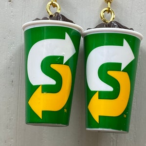 May include: A pair of earrings shaped like a green and white cup with a yellow arrow logo. The logo says "S" with a trademark symbol.