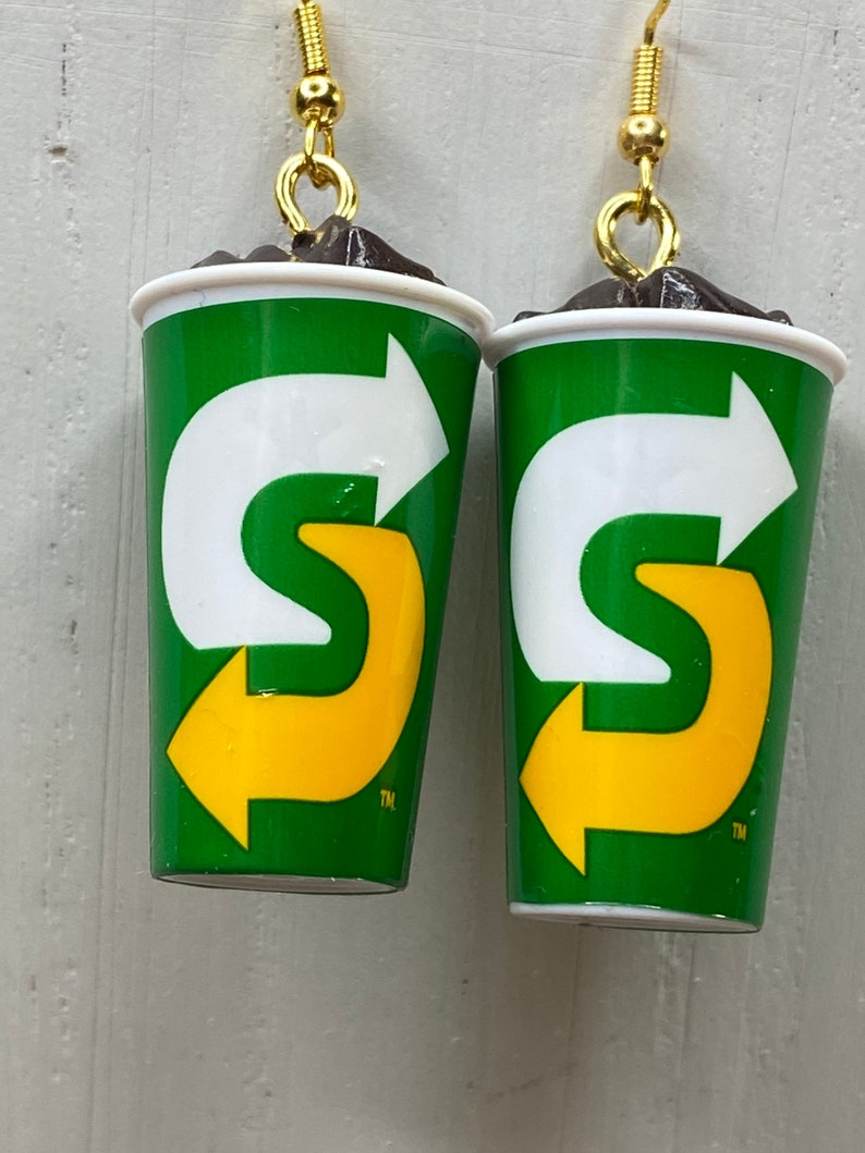 May include: A pair of earrings shaped like a green and white cup with a yellow arrow logo. The logo says "S" with a trademark symbol.