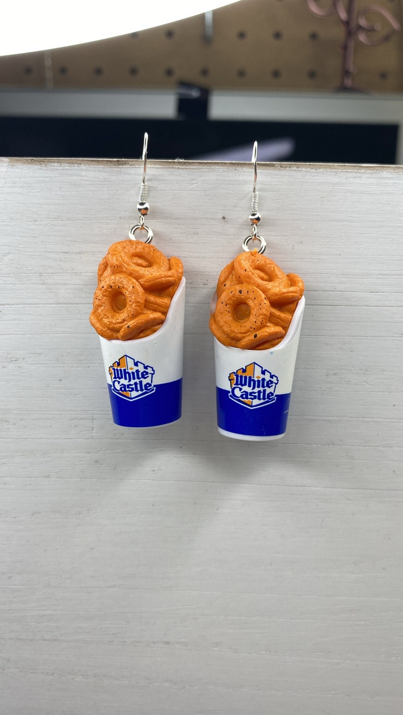 May include: A pair of earrings featuring a White Castle onion ring container with a blue and white logo. The onion rings are orange and have a realistic look.