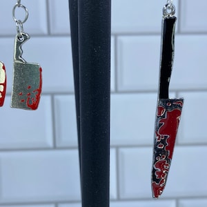 May include: Two pairs of earrings featuring a bloody knife design. One pair is a smaller, more compact knife design, while the other pair is a longer, more detailed knife design.