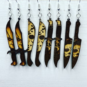 May include: Eight wooden earrings shaped like knives with a wood-burned design. The knives have different designs, including a skull and crossbones, a face, and a simple blade.