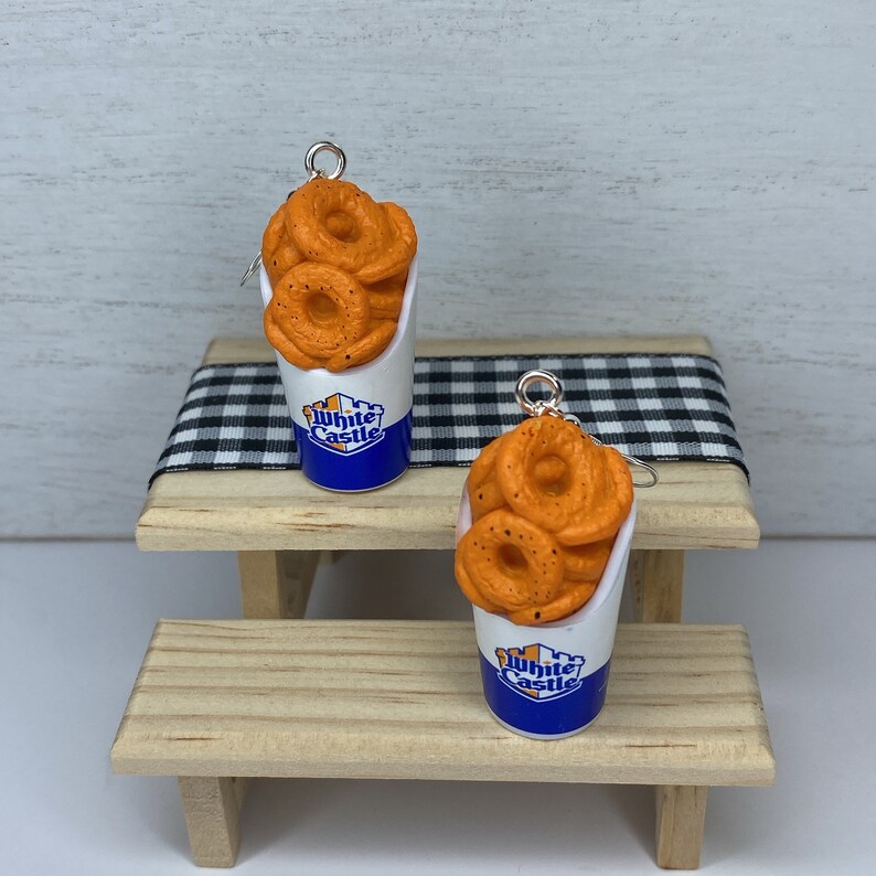 May include: A pair of earrings featuring a White Castle-branded container filled with orange onion rings. The earrings are hanging from a wooden picnic table.