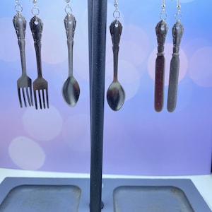 May include: Five pairs of silver-toned earrings hanging on a black stand. The earrings are shaped like forks, spoons, and butter knives.