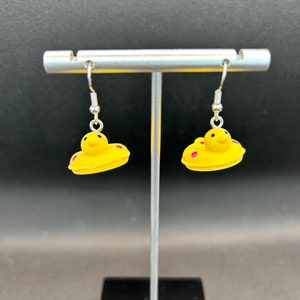 May include: A pair of yellow dangle earrings with pink accents. The earrings are shaped like a cartoon duck with a small, round head and a larger, round body. The duck has a small, pink beak and two small, pink eyes.