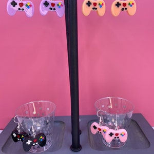 May include: Two pairs of pink and purple video game controller earrings and two pairs of black video game controller earrings displayed on a stand.