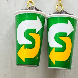 May include: A pair of earrings shaped like green and white cups with a yellow arrow logo. The logo says "S" with an arrow pointing to the right. The earrings have gold hooks.