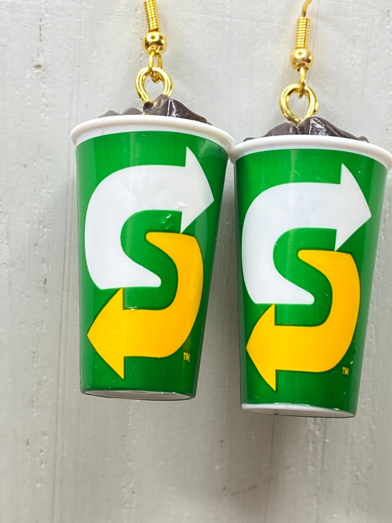 May include: A pair of earrings shaped like green and white cups with a yellow arrow logo. The logo says "S" with an arrow pointing to the right. The earrings have gold hooks.