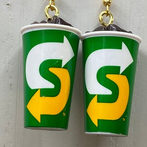 May include: A pair of earrings shaped like a green and white cup with a yellow arrow logo. The logo says "S" with a trademark symbol. The cup is filled with a brown substance that looks like chocolate.