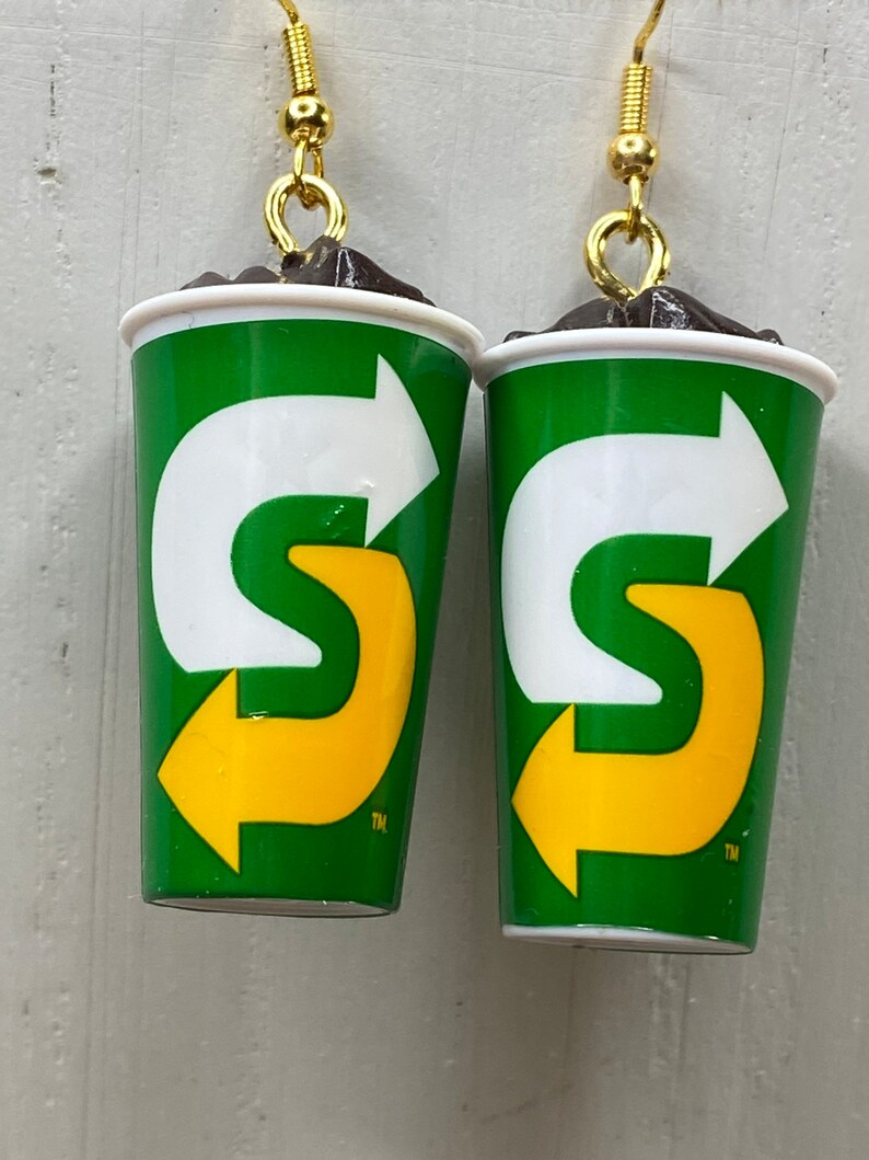 May include: A pair of earrings shaped like a green and white cup with a yellow arrow logo. The logo says "S" with a trademark symbol. The cup is filled with a brown substance that looks like chocolate.