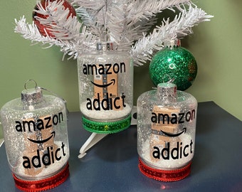 May include: Three glass ornaments with a white glitter background and red ribbon. Each ornament has a black and white image of an Amazon box with the text "amazon addict".