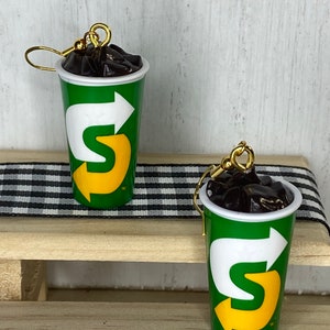 May include: A pair of earrings shaped like a green and yellow cup with a black liquid inside. The cup has a white arrow and the letter "S" in yellow. The earrings are hanging from gold hooks.