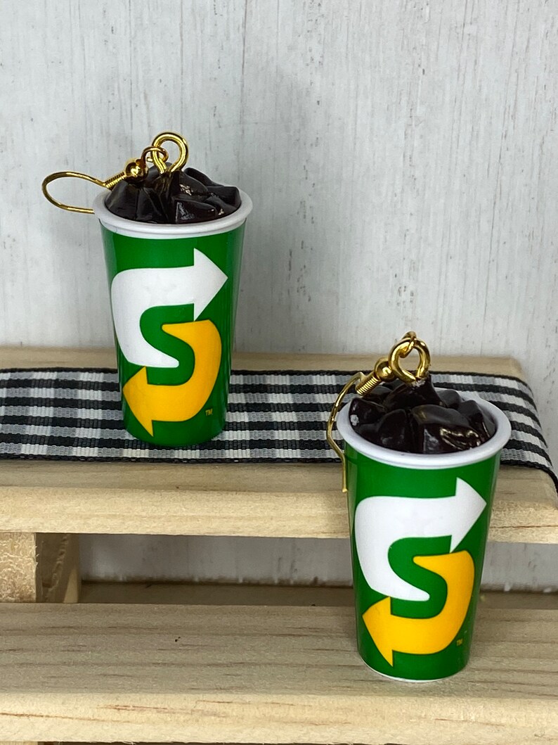 May include: A pair of earrings shaped like a green and yellow cup with a black liquid inside. The cup has a white arrow and the letter "S" in yellow. The earrings are hanging from gold hooks.