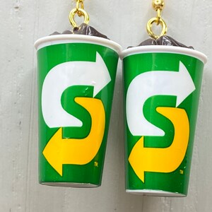 May include: A pair of earrings shaped like a green and yellow 7-Eleven Slurpee cup with a brown top. The cup has a white arrow pointing to the right and a yellow "S" with an arrow pointing to the left. The earrings have gold hooks.