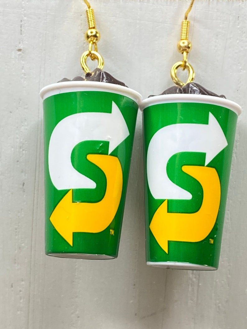 May include: A pair of earrings shaped like a green and yellow 7-Eleven Slurpee cup with a brown top. The cup has a white arrow pointing to the right and a yellow "S" with an arrow pointing to the left. The earrings have gold hooks.