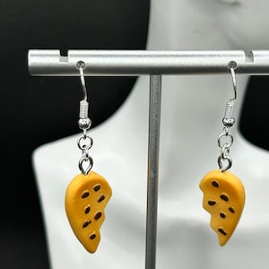 May include: A pair of dangle earrings featuring a yellow, half-circle design with dark brown spots. The earrings are suspended from silver-colored hooks and chains. The earrings are displayed on a white mannequin head.