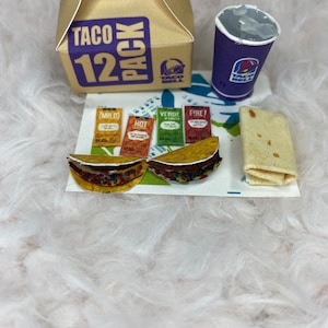 May include: A Taco Bell 12 pack box, two tacos, a purple cup with ice, a burrito, and four packets of Taco Bell sauce: mild, hot, verde, and fire.