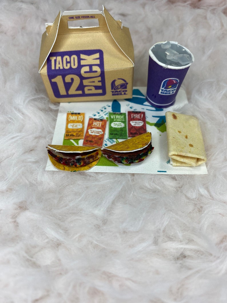 May include: A Taco Bell 12 pack box, two tacos, a purple cup with ice, a burrito, and four packets of Taco Bell sauce: mild, hot, verde, and fire.