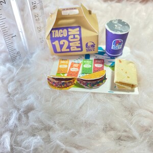 May include: A miniature Taco Bell 12 pack box, a purple cup with the Taco Bell logo, two miniature tacos, a packet of taco seasoning, and a burrito on a white fluffy surface.