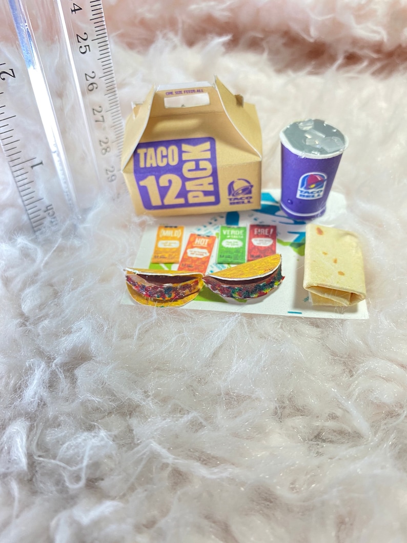 May include: A miniature Taco Bell 12 pack box, a purple cup with the Taco Bell logo, two miniature tacos, a packet of taco seasoning, and a burrito on a white fluffy surface.