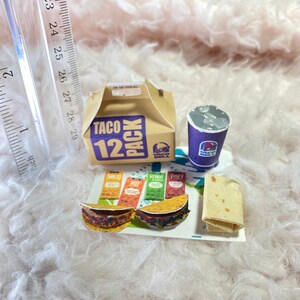May include: A miniature Taco Bell 12 pack box with a purple cup, two miniature tacos, and four packets of sauce. The tacos are on a white paper tray with the text "Taco Bell" printed on it.