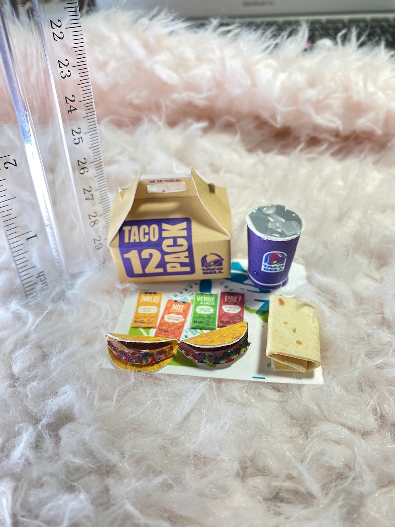May include: A miniature Taco Bell 12 pack box with a purple cup, two miniature tacos, and four packets of sauce. The tacos are on a white paper tray with the text "Taco Bell" printed on it.