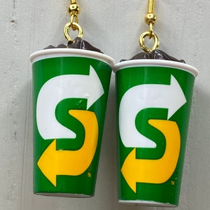 May include: A pair of earrings shaped like a green and white cup with a yellow arrow logo. The logo says "S" with an arrow pointing to the right. The earrings are made of polymer clay and have gold hooks.