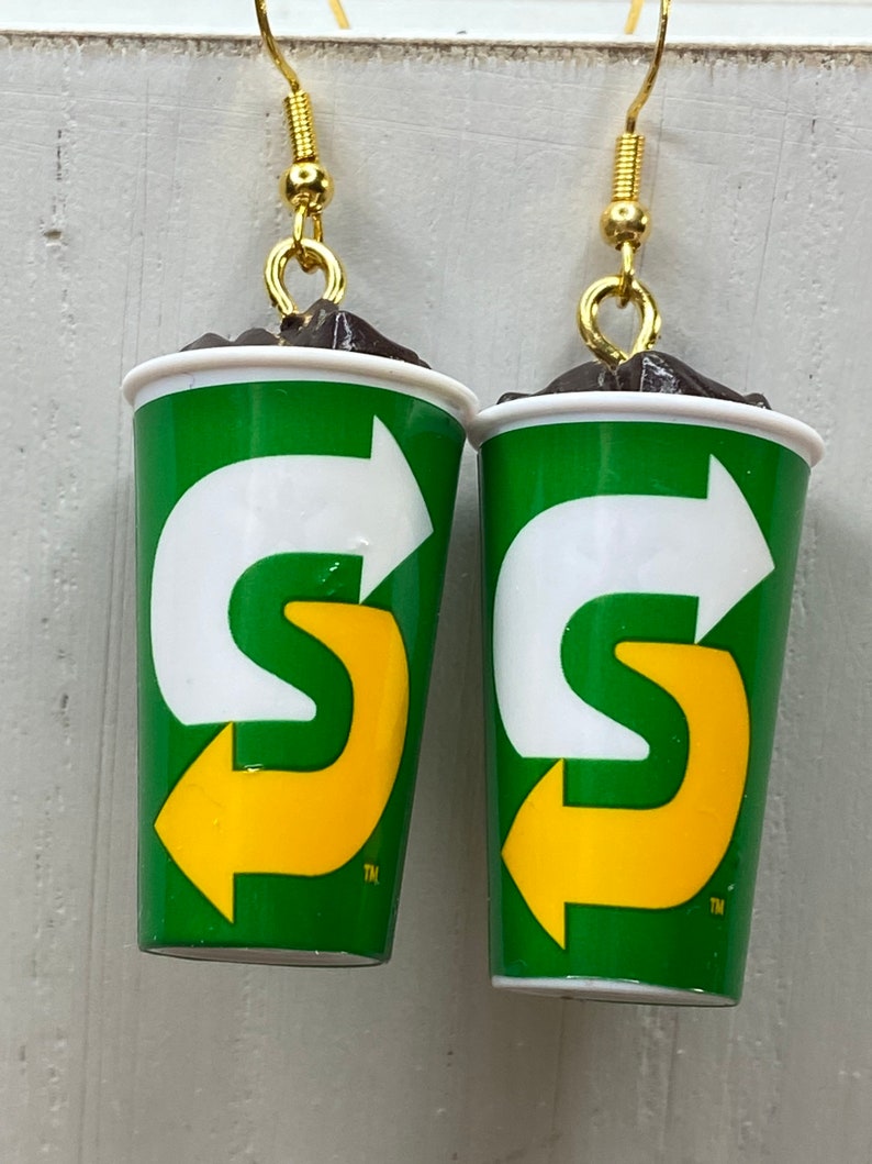 May include: A pair of earrings shaped like a green and white cup with a yellow arrow logo. The logo says "S" with an arrow pointing to the right. The earrings are made of polymer clay and have gold hooks.
