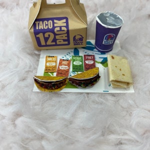 May include: A brown cardboard box with the words "Taco 12 Pack" printed on it. The box is sitting on a white fluffy surface next to a purple cup with the words "Taco Bell" printed on it. There are two small tacos, four packets of hot sauce, and a burrito on a white paper mat.