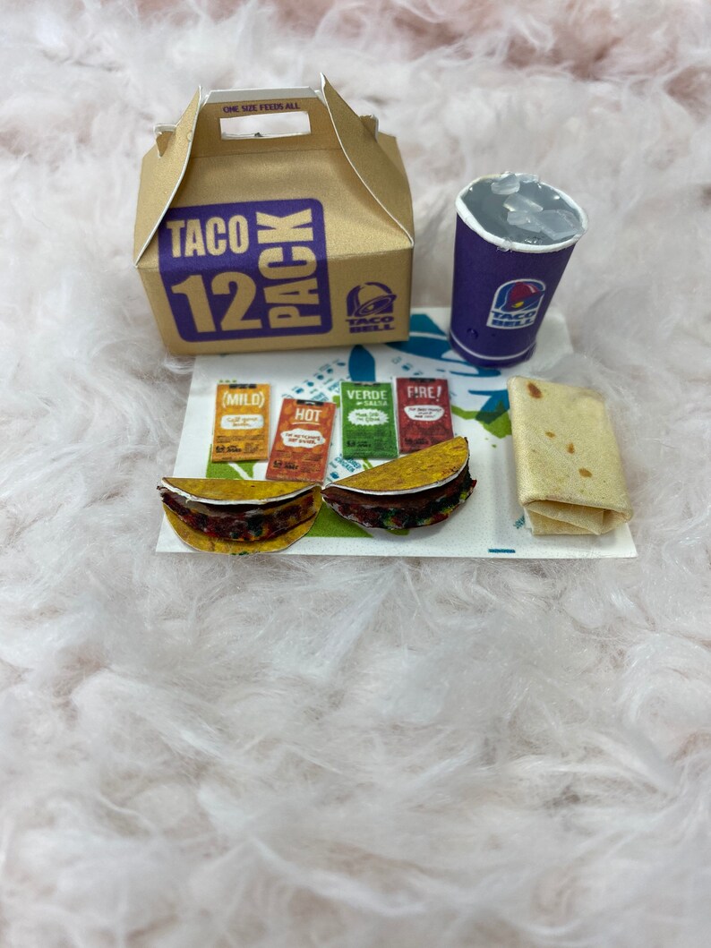 May include: A brown cardboard box with the words "Taco 12 Pack" printed on it. The box is sitting on a white fluffy surface next to a purple cup with the words "Taco Bell" printed on it. There are two small tacos, four packets of hot sauce, and a burrito on a white paper mat.