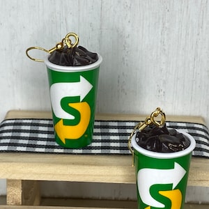 May include: A pair of earrings shaped like a green and yellow cup with a black liquid inside. The cup has a logo with a white arrow and the letter "S" in yellow.