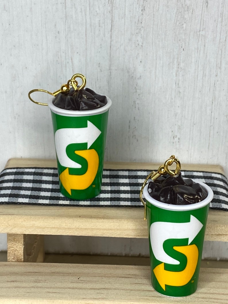 May include: A pair of earrings shaped like a green and yellow cup with a black liquid inside. The cup has a logo with a white arrow and the letter "S" in yellow.
