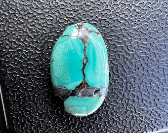 Può includere: Gemma ovale di turchese con venature scure. La pietra lucida mostra una vibrante tonalità verde-azzurro chiaro con motivi irregolari grigio scuro e marroni. La gemma è posta su una superficie nera strutturata.