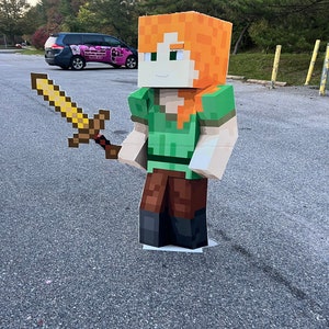 May include: A cardboard cutout of a Minecraft character holding a golden sword. The character has green clothing, brown pants, and orange hair. The cutout is standing on a gray asphalt surface.