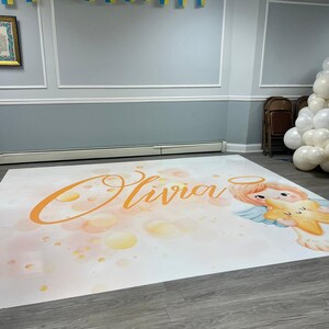 Puede incluir: Un tel&oacute;n de fondo blanco con un efecto acuarela que presenta el nombre "Olivia" en naranja claro y un &aacute;ngel de dibujos animados sosteniendo una estrella dorada. El tel&oacute;n de fondo est&aacute; sobre un suelo de madera.