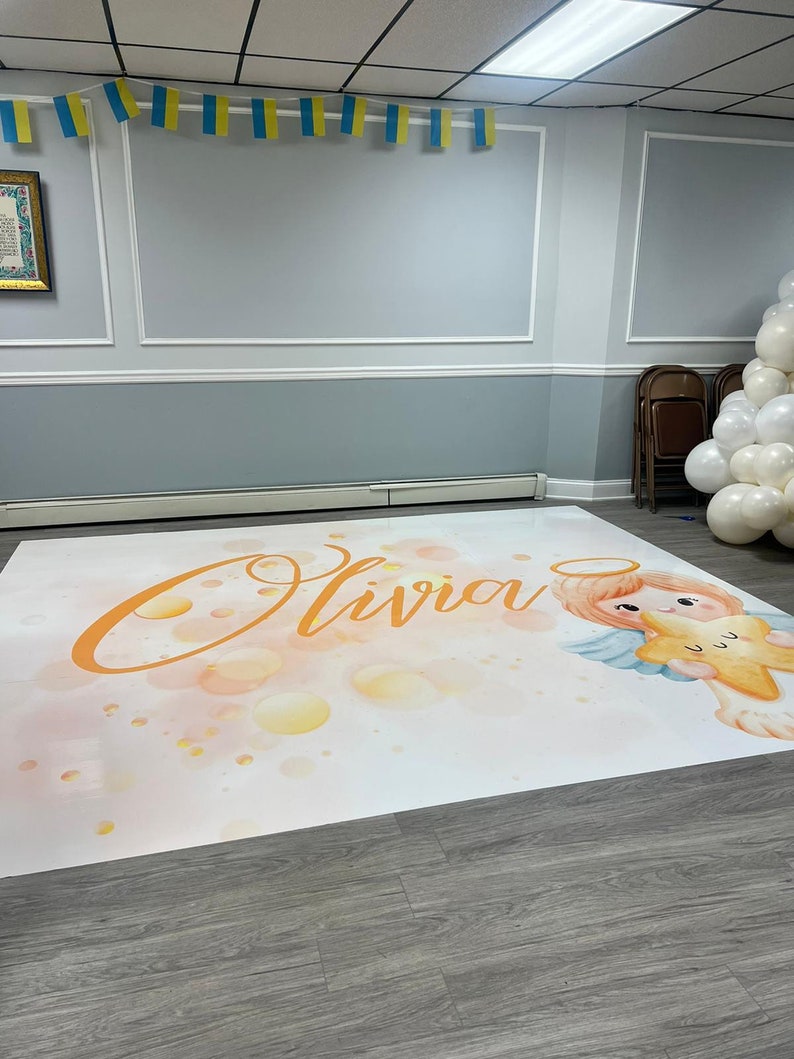 Puede incluir: Un tel&oacute;n de fondo blanco con un efecto acuarela que presenta el nombre "Olivia" en naranja claro y un &aacute;ngel de dibujos animados sosteniendo una estrella dorada. El tel&oacute;n de fondo est&aacute; sobre un suelo de madera.