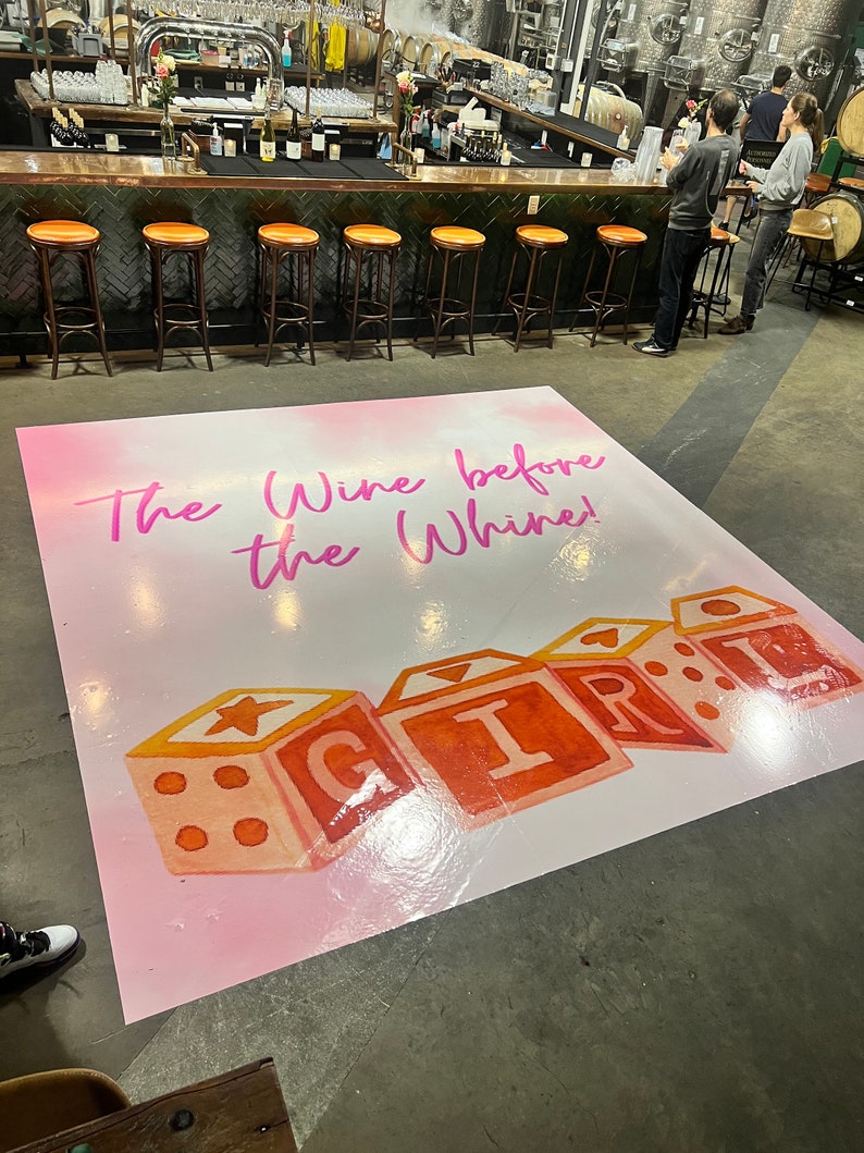 Puede incluir: Una alfombra de suelo rosa y blanca con el texto "The Wine Before The Whine!" y un gr&aacute;fico de bloques de juguetes coloridos que deletrean "GIRL".