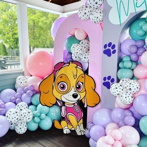 May include: A pink and purple balloon arch with a cardboard cutout of a cartoon dog wearing a pink vest and a blue hat. The arch is decorated with white balloons with paw prints. The text "Mia" is written on a gray background.
