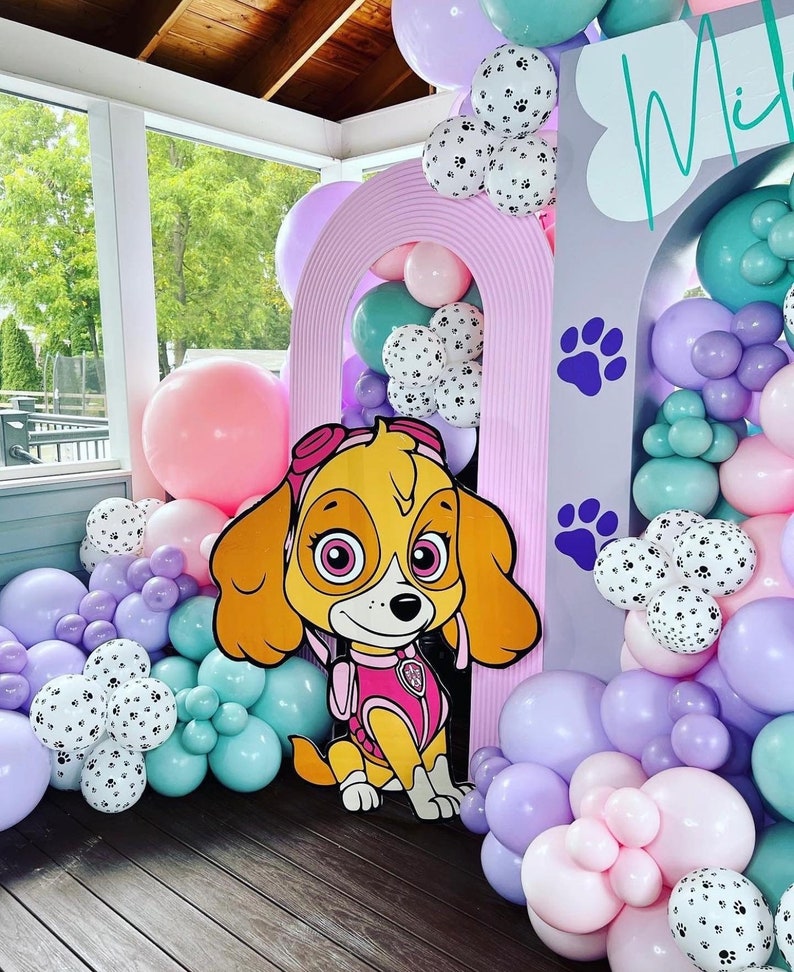 May include: A pink and purple balloon arch with a cardboard cutout of a cartoon dog wearing a pink vest and a blue hat. The arch is decorated with white balloons with paw prints. The text "Mia" is written on a gray background.