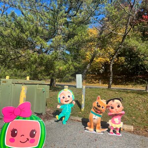 May include: A set of four cardboard stand-up figures featuring characters from the children's show CoComelon. The figures include a pink watermelon character, a blue and green character wearing headphones, a brown dog, and a pink and yellow character with a pink bow.