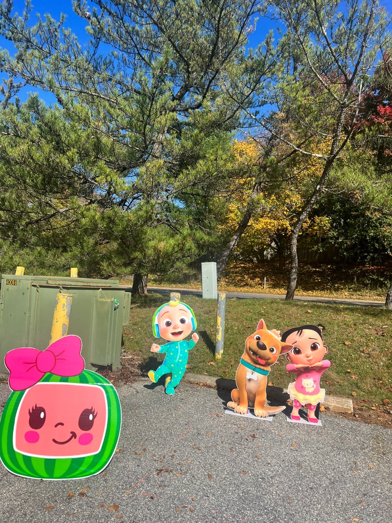 May include: A set of four cardboard stand-up figures featuring characters from the children's show CoComelon. The figures include a pink watermelon character, a blue and green character wearing headphones, a brown dog, and a pink and yellow character with a pink bow.