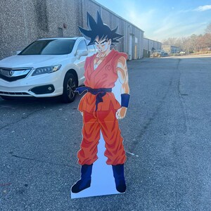 May include: A cardboard cutout of a cartoon character wearing an orange jumpsuit with a black belt and black boots. The character has black hair and a serious expression.