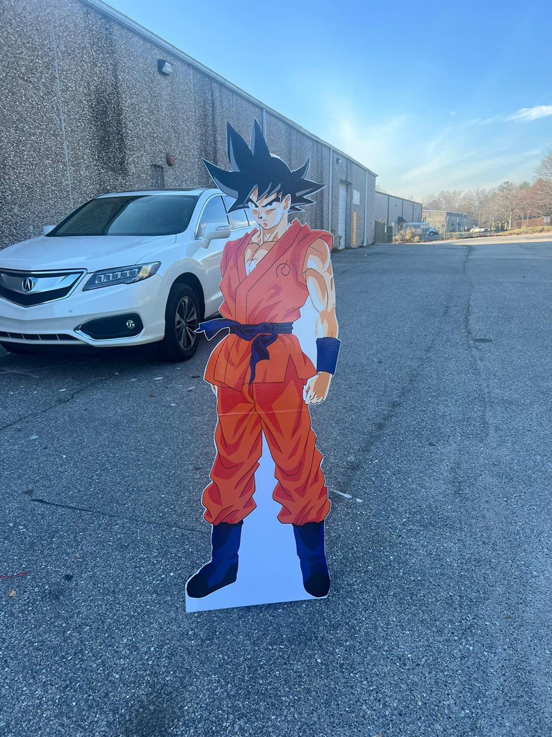 May include: A cardboard cutout of a cartoon character wearing an orange jumpsuit with a black belt and black boots. The character has black hair and a serious expression.