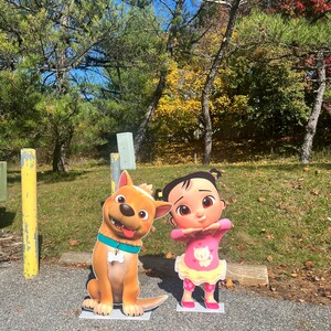May include: A brown and white cartoon dog with a blue collar and a white bone charm stands next to a cartoon girl with pink hair, wearing a pink shirt with a white cat graphic and a pink skirt. The characters are standing on a gray asphalt surface.