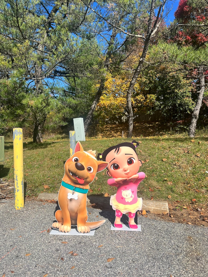 May include: A brown and white cartoon dog with a blue collar and a white bone charm stands next to a cartoon girl with pink hair, wearing a pink shirt with a white cat graphic and a pink skirt. The characters are standing on a gray asphalt surface.