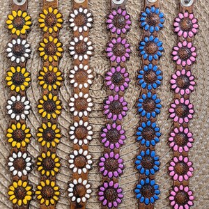 Op de afbeelding: Zes leren armbanden met zonnebloemmotieven. De armbanden zijn bruin met verschillende kleuren voor het midden van de zonnebloemen: geel, wit, roze, blauw en lichtroze.