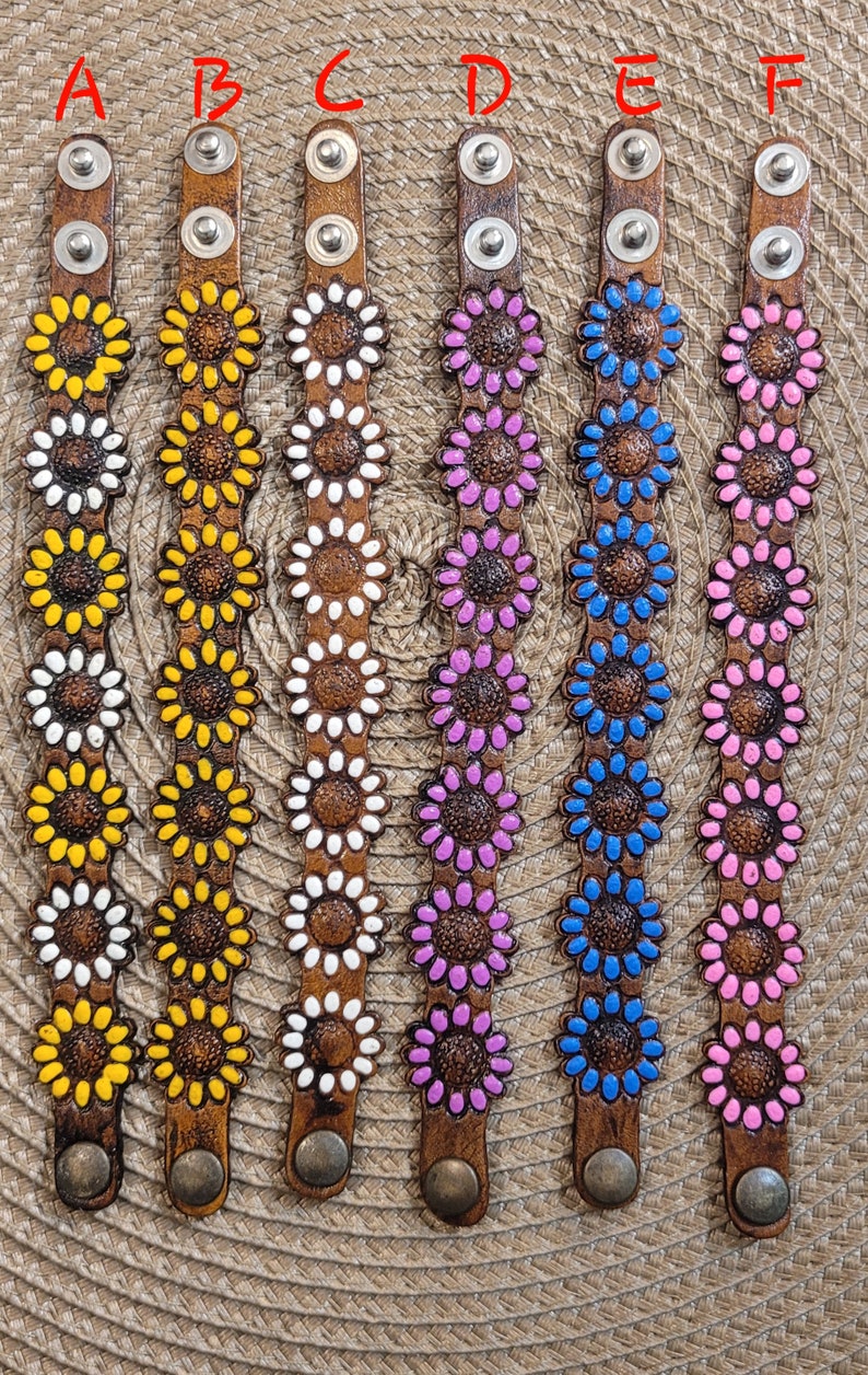 Op de afbeelding: Zes leren armbanden met zonnebloemmotieven. De armbanden zijn bruin met verschillende kleuren voor het midden van de zonnebloemen: geel, wit, roze, blauw en lichtroze.