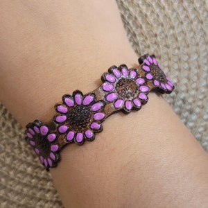 Op de afbeelding: Een bruin leren armband met paarse bloemenaccenten. De armband is versierd met meerdere bloemendesigns.