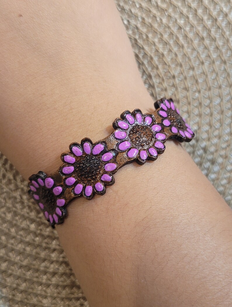 Op de afbeelding: Een bruin leren armband met paarse bloemenaccenten. De armband is versierd met meerdere bloemendesigns.